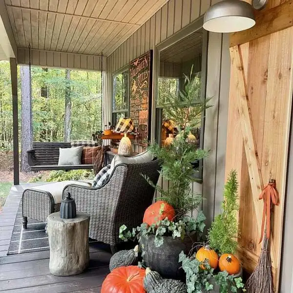 Wood-paneled siding, a wicker chair, plaid pillows, rustic Halloween décor and potted evergreens with pumpkins on a forest-side porch