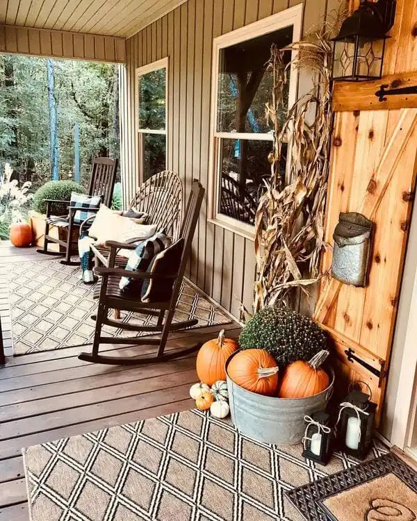 A rustic porch outfitted with rocking chairs, layered rugs, plaid pillows, corn stalks and a galvanized tub filled with pumpkins and mums.