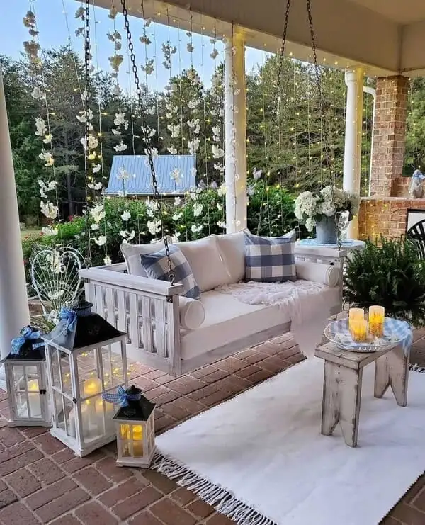A dreamy porch featuring a white hanging daybed, blue gingham pillows, overflowing floral garlands, flickering lanterns, and a white fringed rug.
