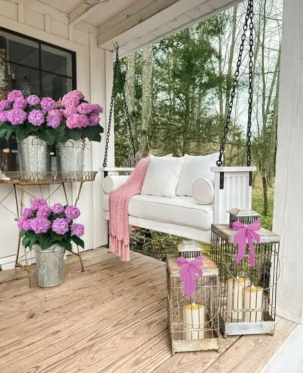 A farmhouse-style porch with a white swing, galvanized planters of pink hydrangeas, wire lanterns with bows and a blush throw blanket.