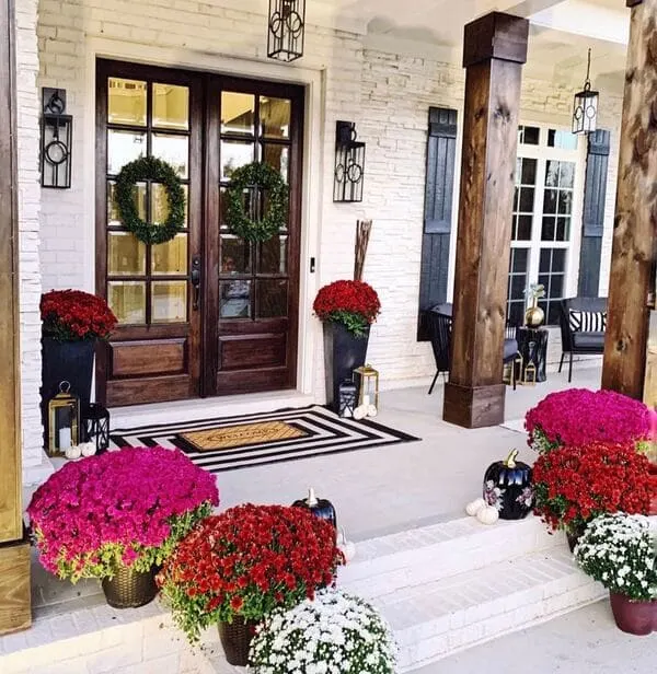 A brick front porch with rich wood doors, fall mums in vibrant kinds, tall black planters, and tiered lanterns.