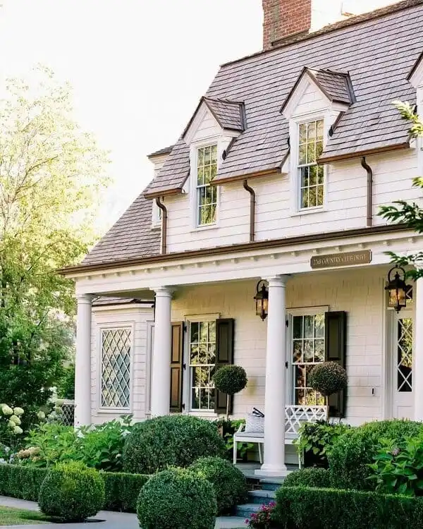 A classic colonial-style house featuring white columns, diamond-pane windows, black shutters, and sculpted greenery on a traditional front porch.