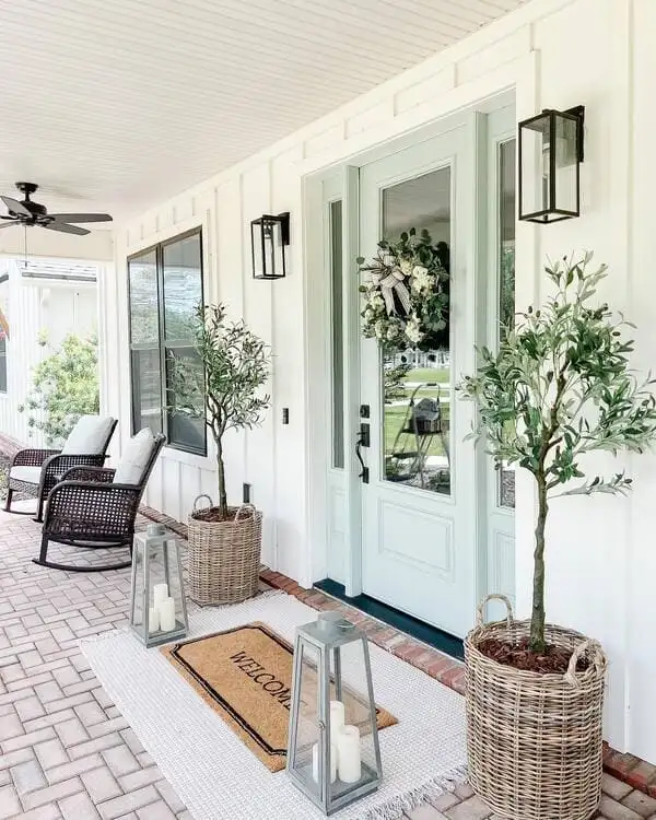 A farmhouse-style porch with board-and-batten siding, mint green front doors, wicker planters holding olive trees and oversized candle lanterns.