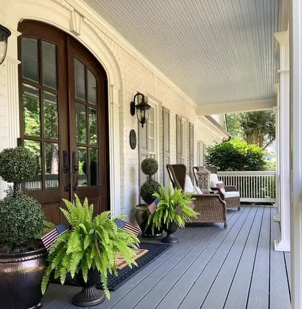 A Southern-style porch with pale blue ceiling, double dark doors, wicker chairs, ferns, and topiary planters.