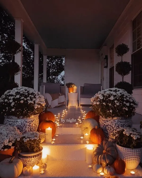 A moody fall porch illuminated with candlelight, pumpkins, mums in chinoiserie planters and soft seating at the far end.