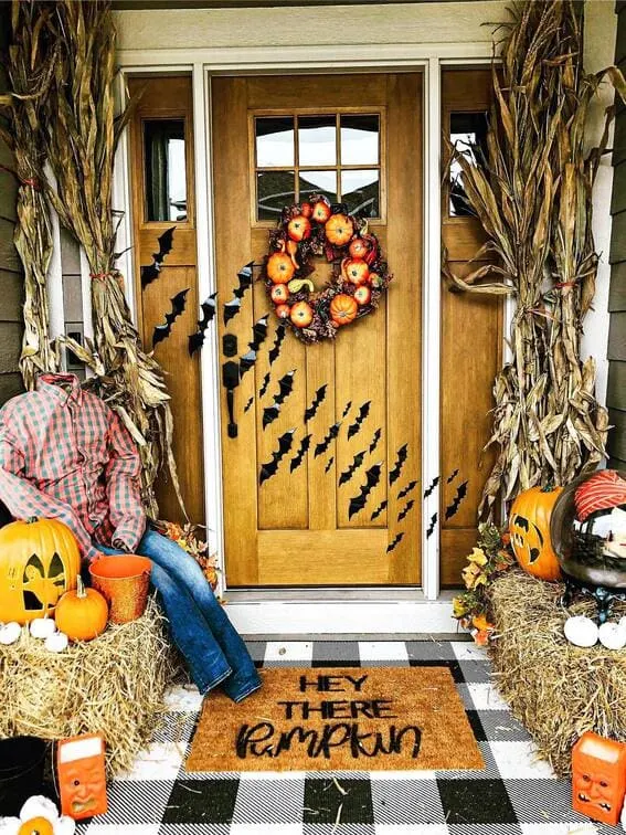 This porch combines spooky with harvest charm, featuring black bats flying up the wooden door and a headless scarecrow lounging on a hay bale.