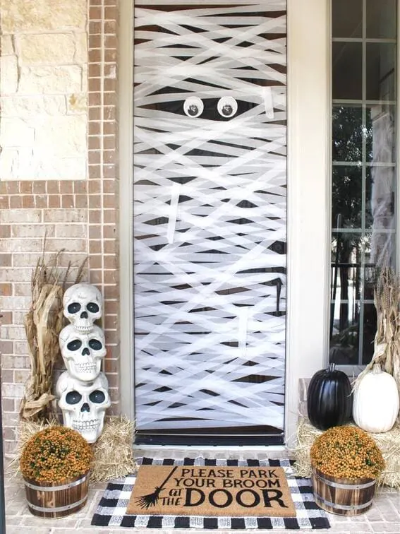 This porch features a playful mummy door design, where the door is wrapped in white streamers, resembling a mummy with large googly eyes peeking through.