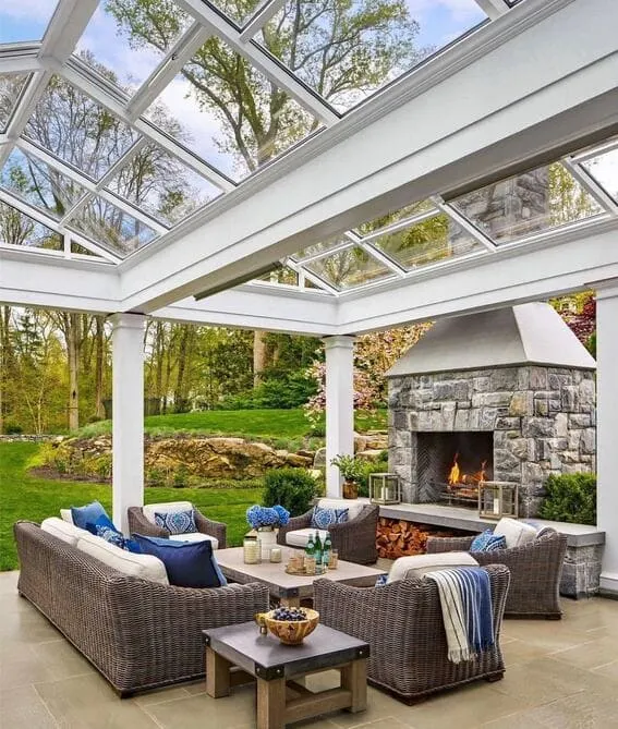 Beautiful patio with the fireplace of a stone under a glass pergola to give an airy yet cozy appearance.
