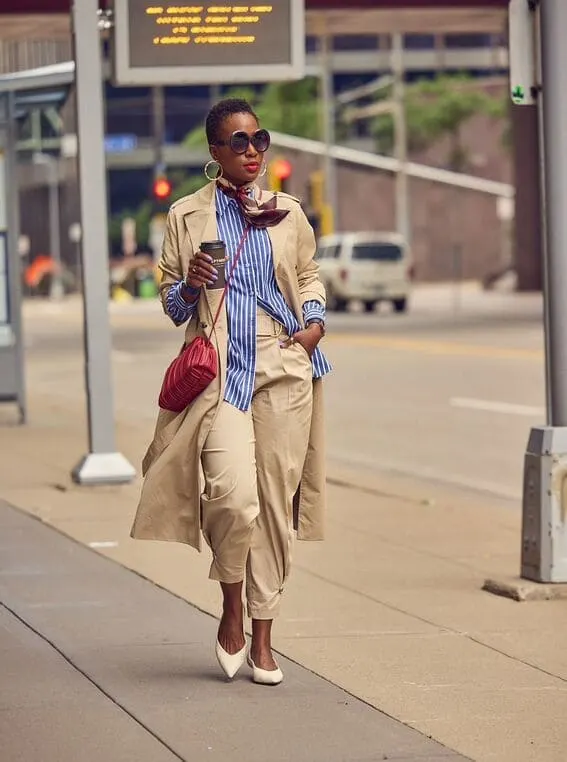 An elegant woman is styled in a blue-striped oversized shirt paired with a trench coat, tailored trousers, and bold accessories.