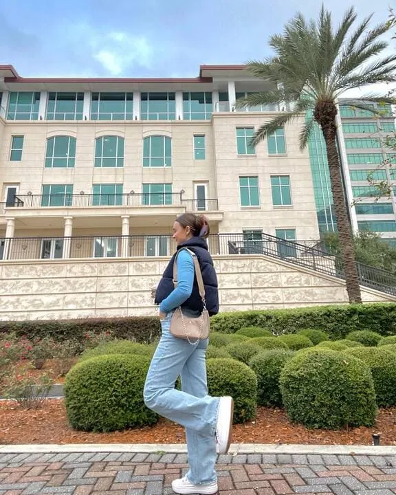 A woman wearing a navy puffer vest, light blue long-sleeve top, light-wash flare jeans, and white sneakers, accessorized with a beige crossbody bag.