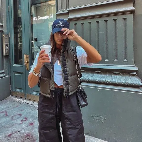 A woman wearing an olive puffer vest, white cropped tee, black wide-leg pants, and a baseball cap, while holding a coffee cup.