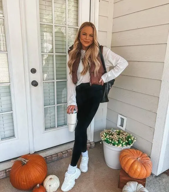 A woman is wearing a brown puffer vest, a white long-sleeve top, black leggings, white sneakers, and a tumbler.