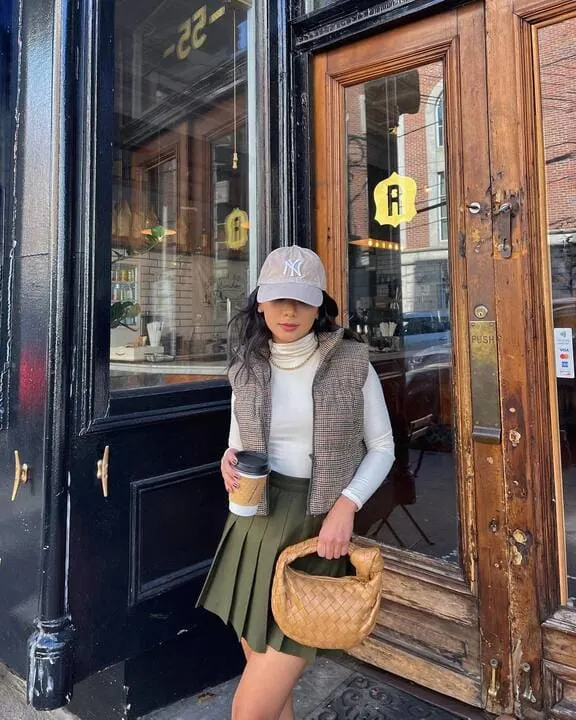 A woman wearing a plaid puffer vest, white turtleneck, olive pleated skirt, and a baseball cap while holding a woven handbag and coffee cup.