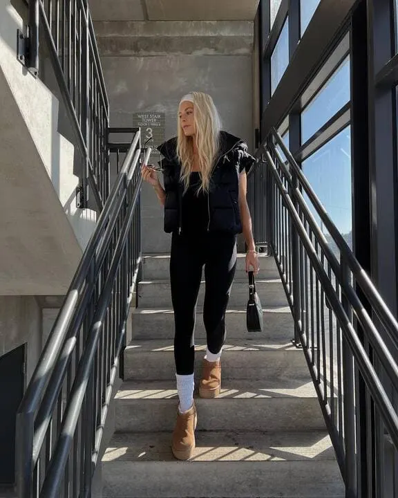 A woman wearing a cropped black puffer vest, black leggings, white socks, platform slippers, and a headband while carrying a small handbag.