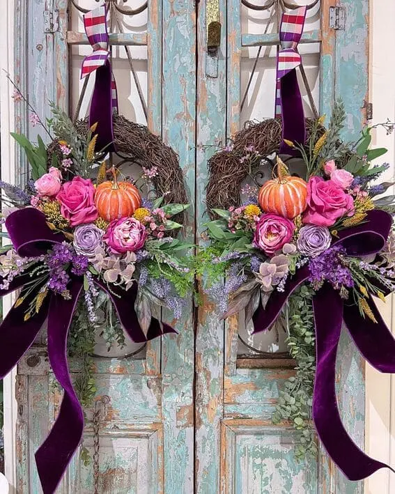 A pair of stunning wreaths adorned with vibrant orange pumpkins, lush florals, and rich velvet ribbons in deep purple tones.