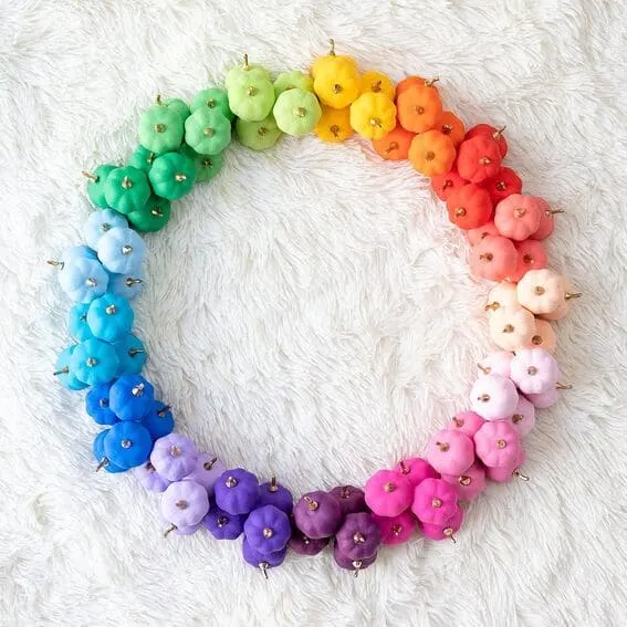 A vibrant, colorful wreath featuring mini pumpkins arranged in a stunning rainbow gradient, accented with gold stems.