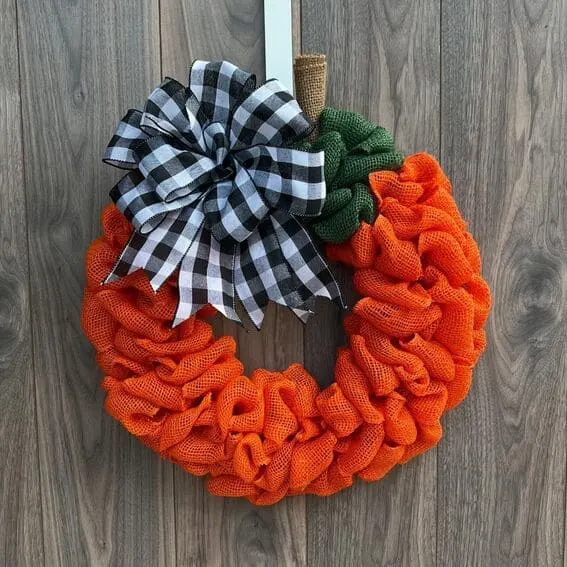 A vibrant pumpkin-shaped wreath made of orange and green burlap, accented with a bold black-and-white gingham bow.