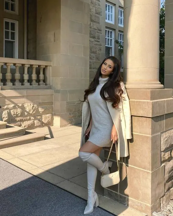 A woman is wearing an white turtleneck sweater worn with long coat and over-the-knee boots.